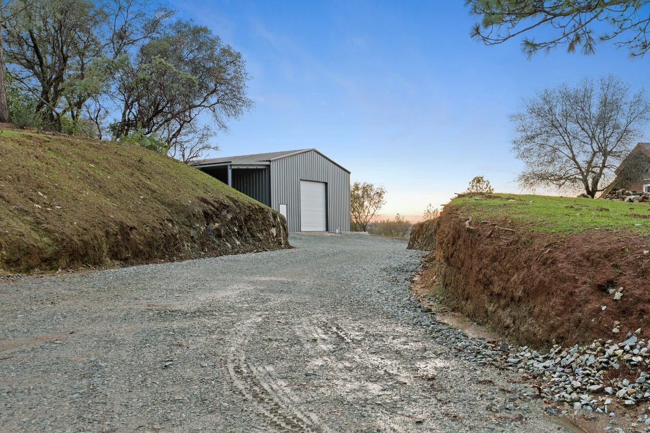 Gravel driveway curving up a grassy hillside toward a steel shop building